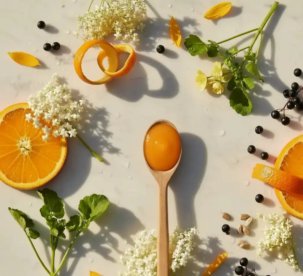 Spoon with orange substance surrounded by oranges, flowers, and berries on a light surface
